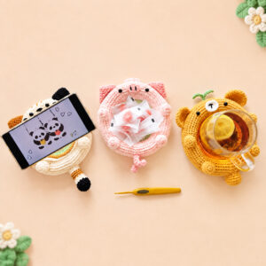Three crocheted animal dishes side by side on a peach background: a cream cat with one black ear and one brown ear propping up a phone, a pink pig holding a handful of wrapped sweets, and a caramel bear with two green leaves on its head and a glass mug of lemon tea standing down inside it, with a yellow crochet hook in front