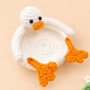 A crocheted white duck coaster seen from above on a peach background: the duck lies on his back as a round dish with a raised rim, his round white head with two shiny black eyes and an orange beak resting on one edge, a long white arm curving along the rim on each side, and two orange webbed feet sticking out over the far edge