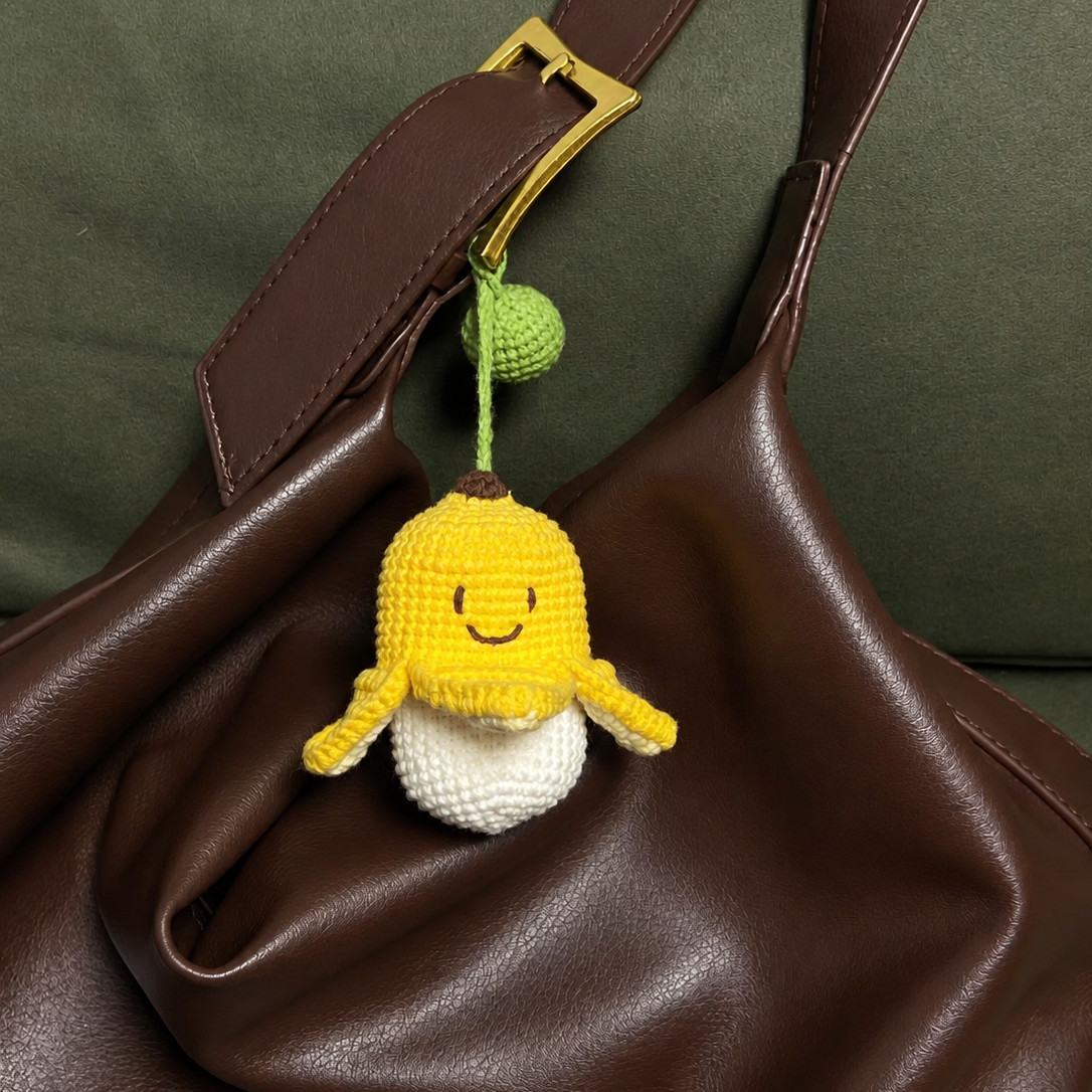A crocheted banana key pouch hanging by its green strap and green ball from the gold buckle of a dark brown leather bag: a smiling yellow peel with a dark brown tip and four flaps turned out over a cream pouch