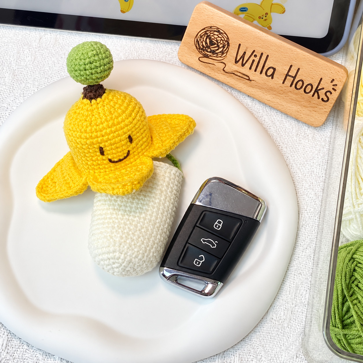 A crocheted banana key pouch lying on a white tray beside a black car key fob and a wooden Willa Hooks sign: the smiling yellow peel slid up with its flaps turned out, the long cream pouch below it, and a green ball on top