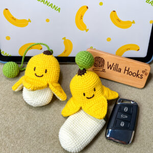 Two crocheted banana key pouches on a grey carpet in front of a tablet showing a banana wallpaper: the one on the left has its yellow peel pulled down over a cream pouch, its four flaps turned out and a green ball on a green strap; the one on the right has its peel slid up so the long cream pouch shows, with a green ball on top, beside a black car key fob and a wooden Willa Hooks sign