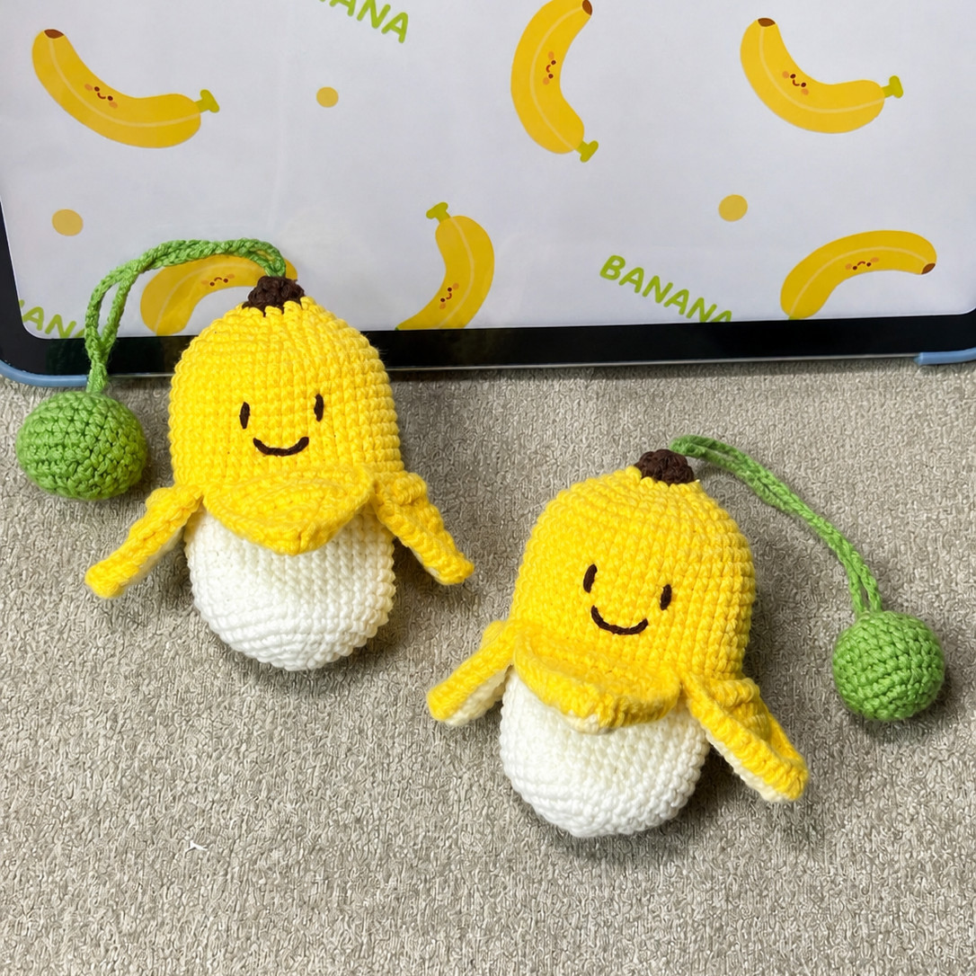 Two crocheted banana key pouches on a grey carpet in front of a tablet showing a banana wallpaper, each a smiling yellow peel with a dark brown tip and four flaps turned out over a cream pouch, with a green ball on a green strap