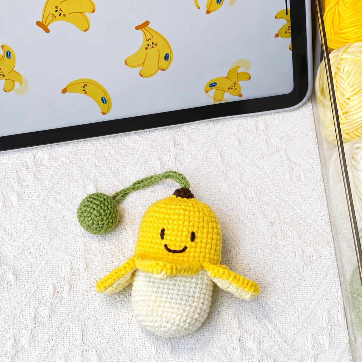 A crocheted banana key pouch lying on a white textured cloth in front of a tablet showing a banana wallpaper: a smiling yellow peel with a dark brown tip, its four flaps turned out over a cream pouch, and a green strap running to a green ball, with balls of yellow and pale yellow yarn to one side
