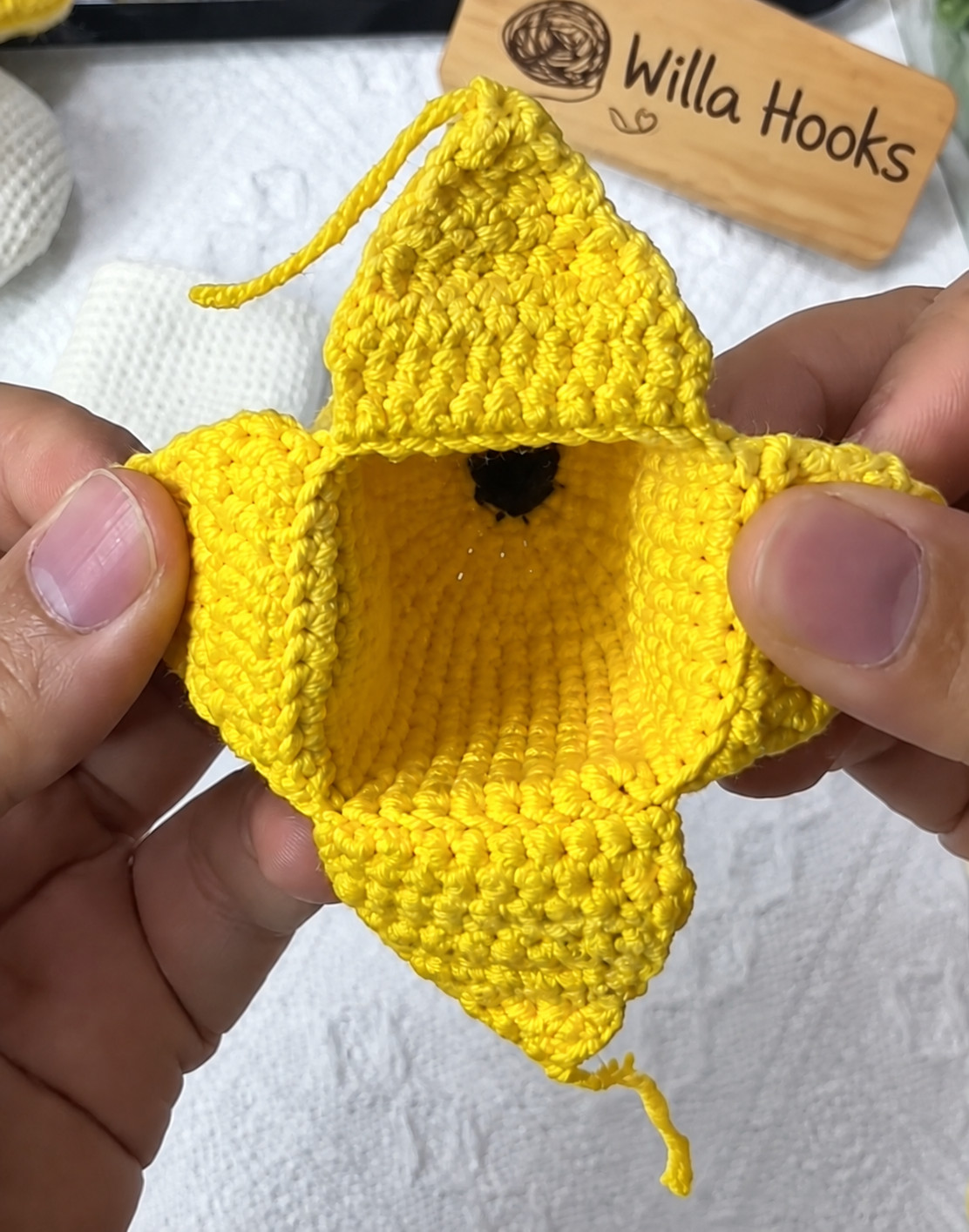 The yellow crocheted peel of the banana key pouch before its linings go on, held open in two hands: four flaps opened out round a square opening, looking down into the cup to its dark brown tip, with a wooden Willa Hooks sign behind