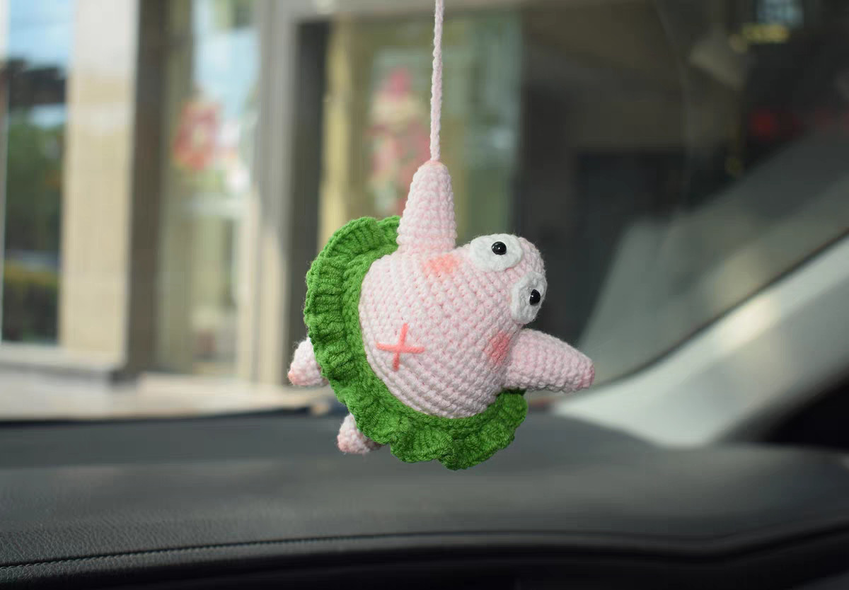 A crocheted pink starfish hanging from a pink cord in front of a window, turned to one side: white eyes with black pupils, pink cheeks, a pink cross on its tummy, one arm out, and a green ruffle skirt