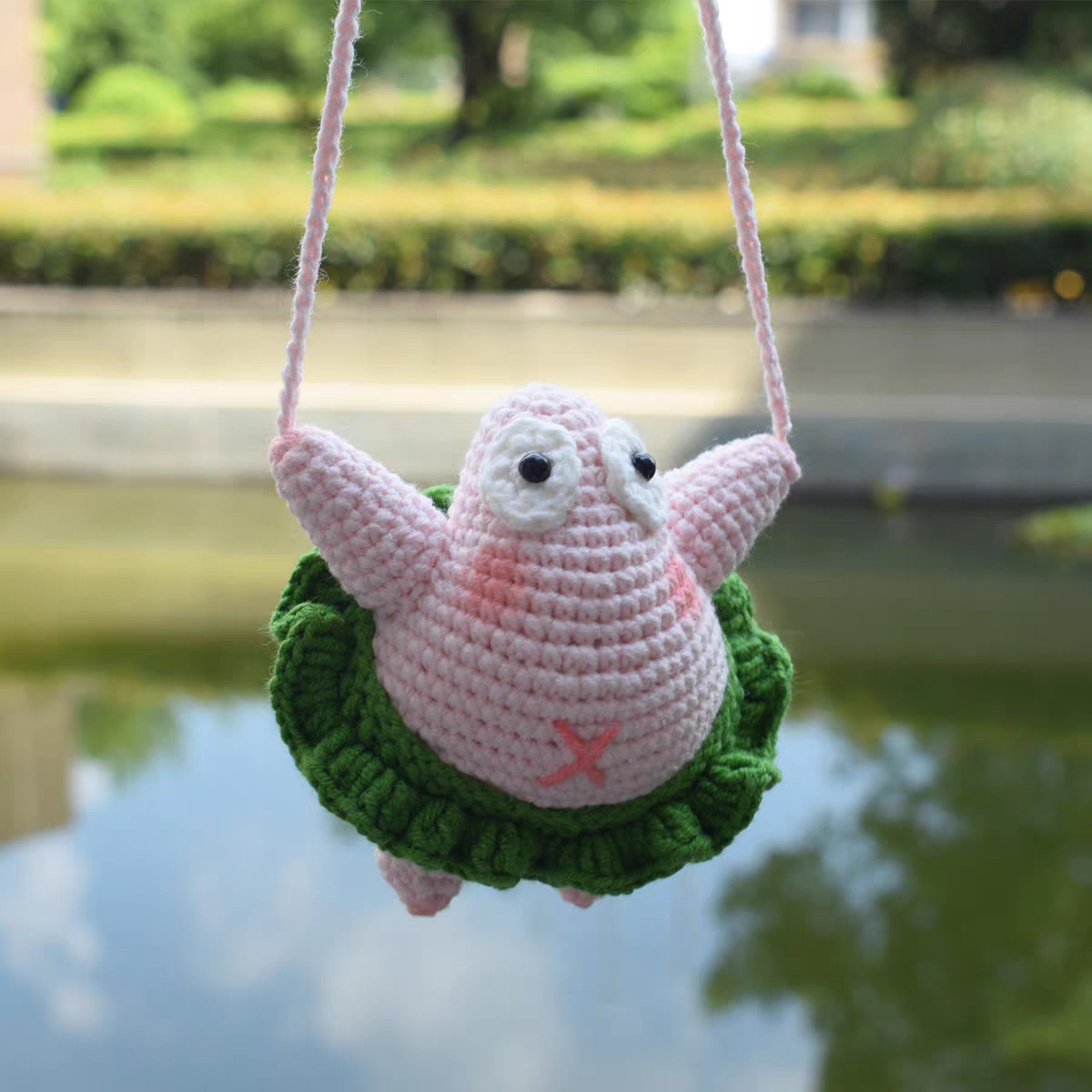A crocheted pink starfish sitting in a loop of pink cord that runs through the tips of both raised arms, above water: white eyes with black pupils, pink cheeks, a pink cross on its tummy, a green ruffle skirt and two stubby legs