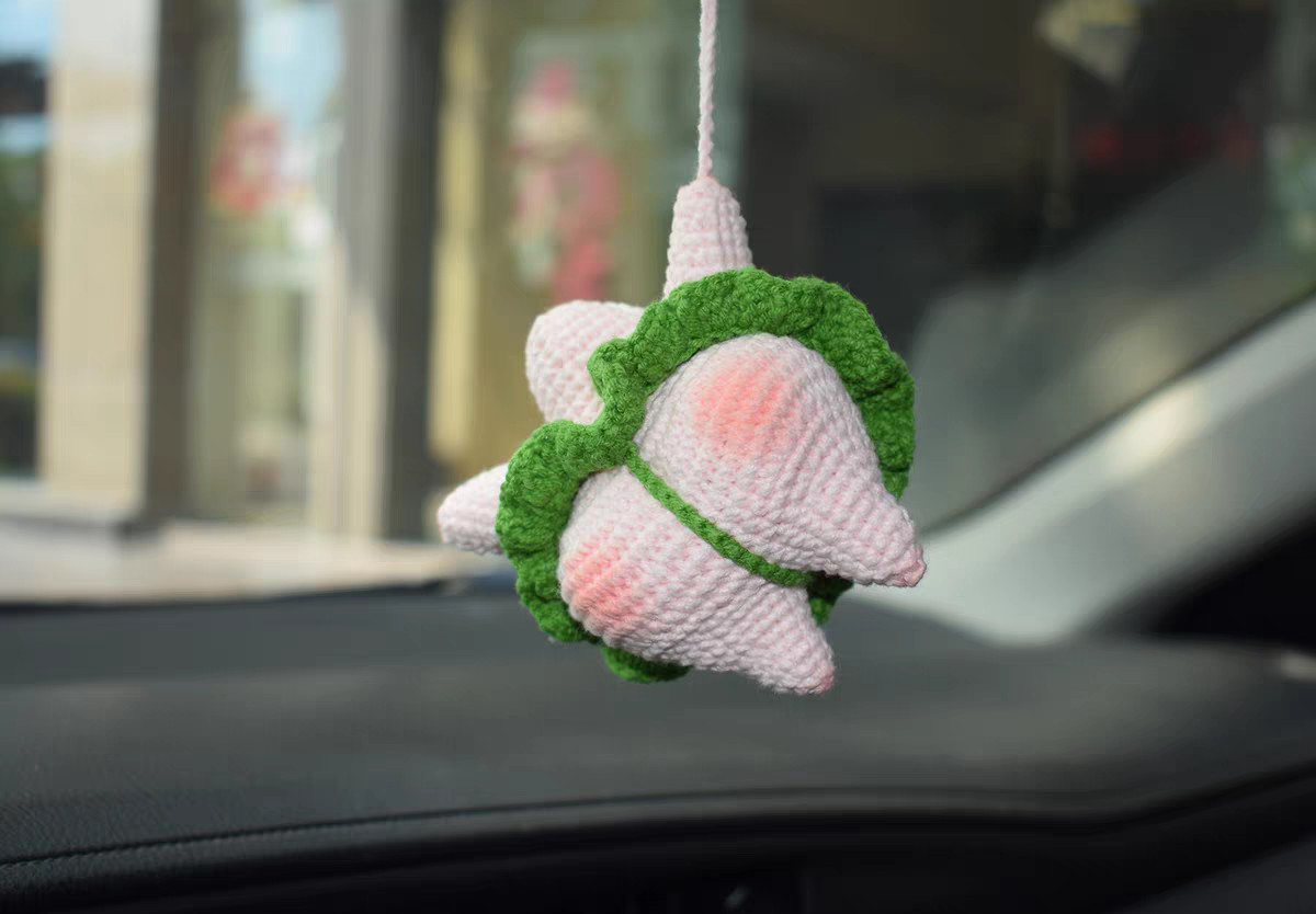 The back of a crocheted pink starfish hanging from a pink cord at its top point: the raised seat with pink blush on both sides, a green strap running between the two legs, and the ruffle skirt round its waist