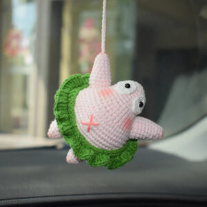 A crocheted pink starfish hanging from a pink cord tied to its top point: white eyes with black pupils, pink cheeks, a pink cross on its tummy, one arm pointing out, and a green ruffle skirt round its middle