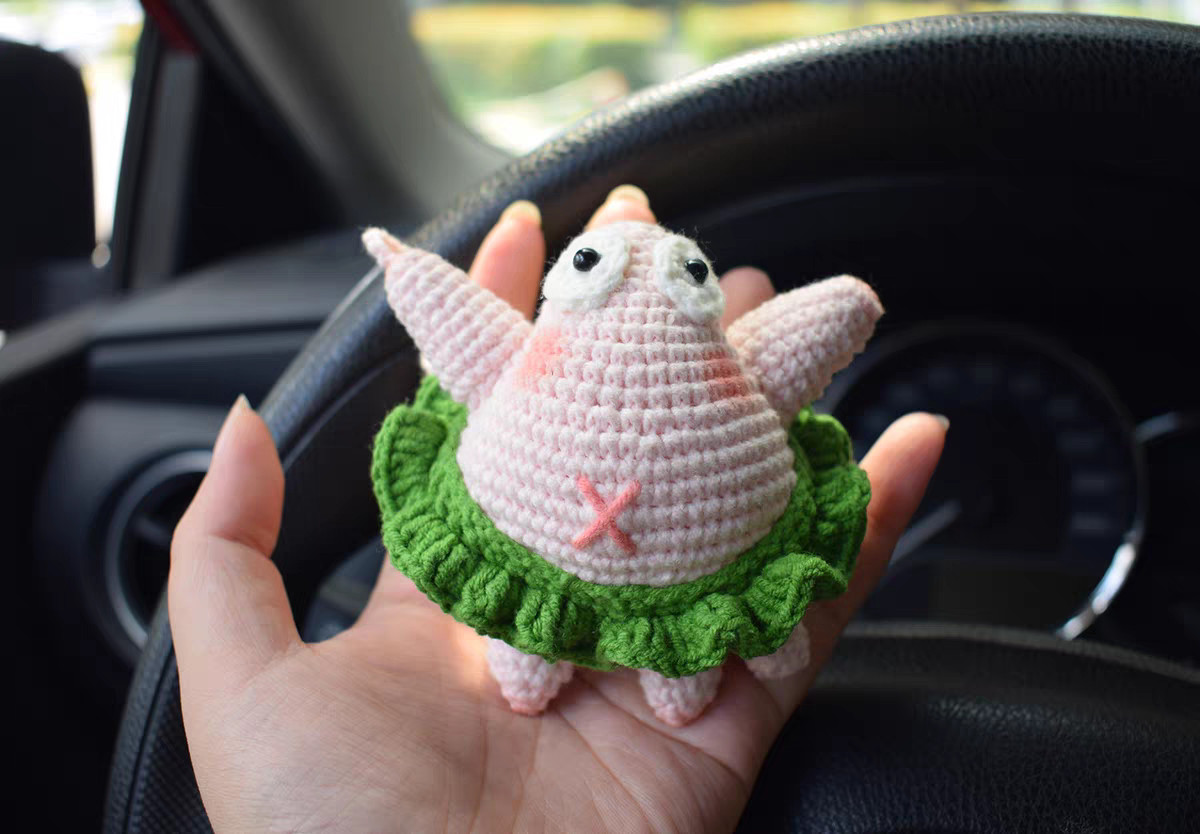 A crocheted pink starfish standing on an open hand, seen straight on: white eyes with black pupils at the top, both arms raised at the sides, pink cheeks, a pink cross on its tummy, a green ruffle skirt and two stubby legs below