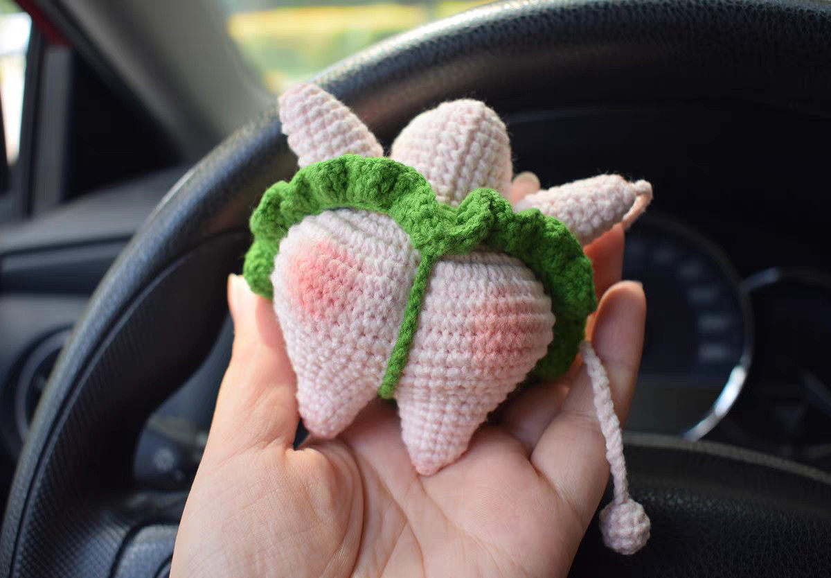 The back of a crocheted pink starfish held in a hand: the rounded, raised seat with pink blush on each side, a green strap running from the ruffle skirt down between the two legs, and a pink cord ending in a small crocheted ball