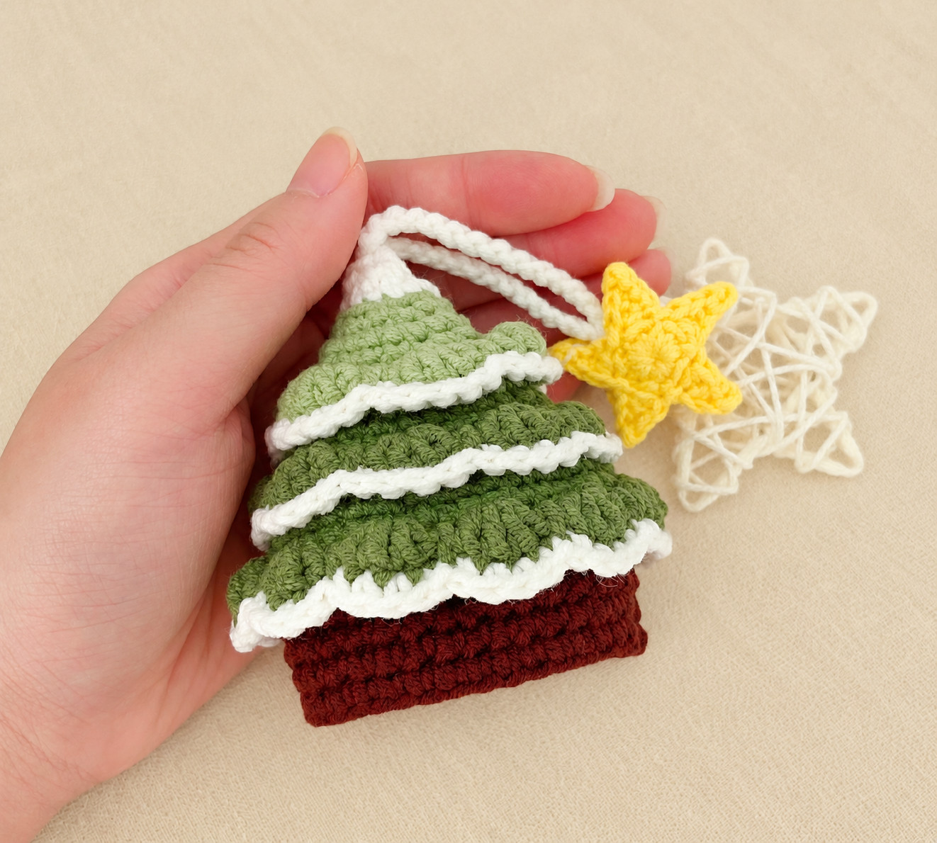 A crocheted Christmas tree key pouch lying in an open hand on a beige cloth: three tiers of green leaves edged in cream over a dark red base, its cream cord looped out of the top to a yellow crocheted star, beside a white rattan star
