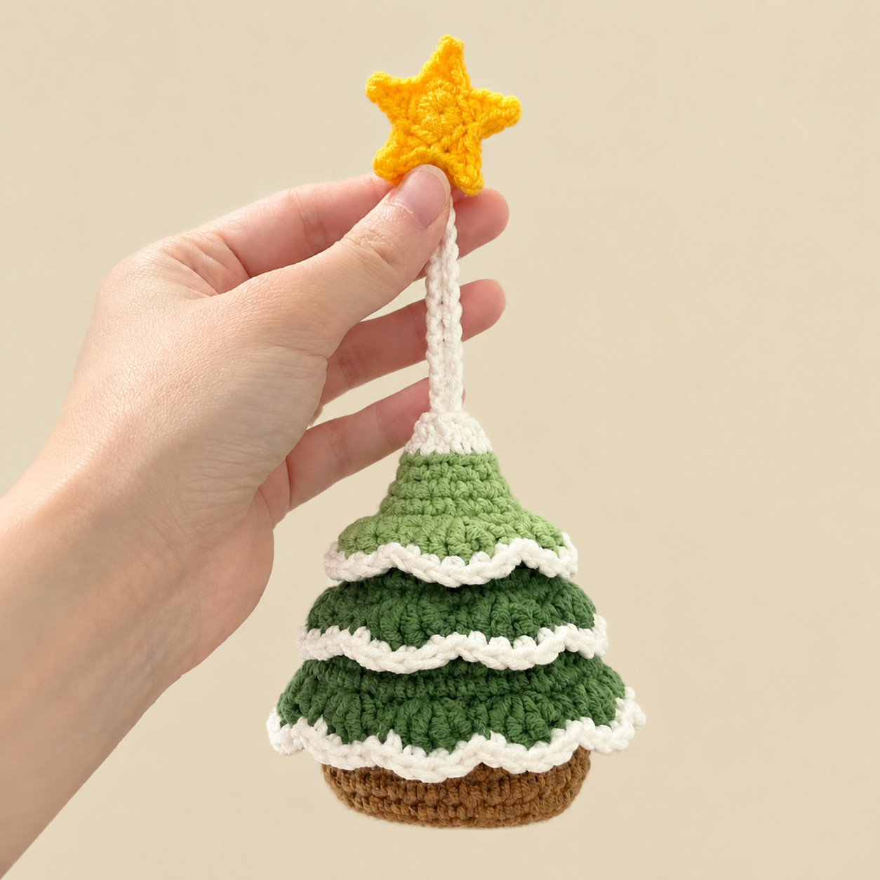 A crocheted Christmas tree key pouch hanging from its yellow star, held between finger and thumb against a cream background: a cream cord, a cream tip, three tiers of green scalloped leaves edged in cream, and a brown base