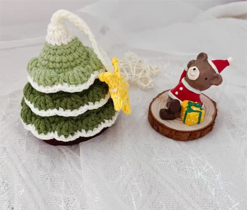 A crocheted Christmas tree key pouch on a sheer white cloth, its cream cord looped over the top to a yellow crocheted star at its side, beside a small figure of a bear in a red Santa hat and coat with a yellow gift, sitting on a slice of wood