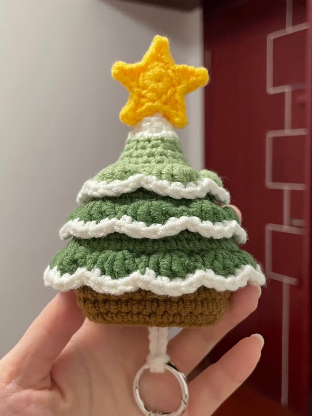 A crocheted Christmas tree key pouch held in one hand in front of a dark red wall: a yellow star on top, three tiers of green leaves edged in cream, a brown base, and a cream cord with a metal ring hanging below it