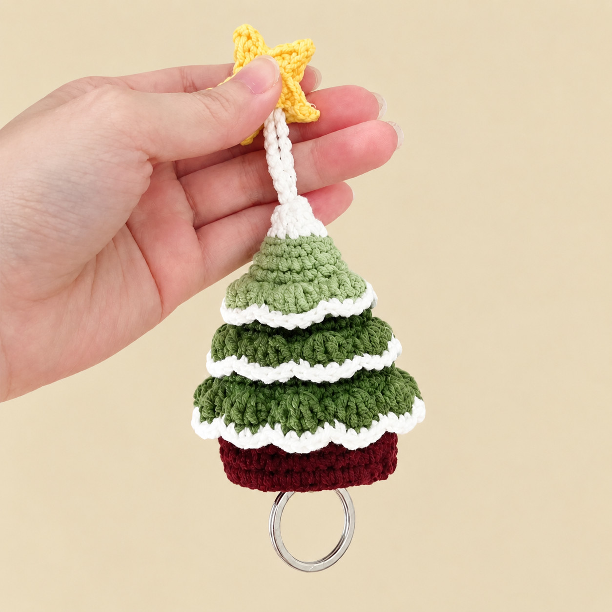 A crocheted Christmas tree key pouch held up by its yellow star against a cream background: a cream cord running down to a cream tip, three tiers of green scalloped leaves edged in cream, a dark red base, and a metal key ring hanging just below it