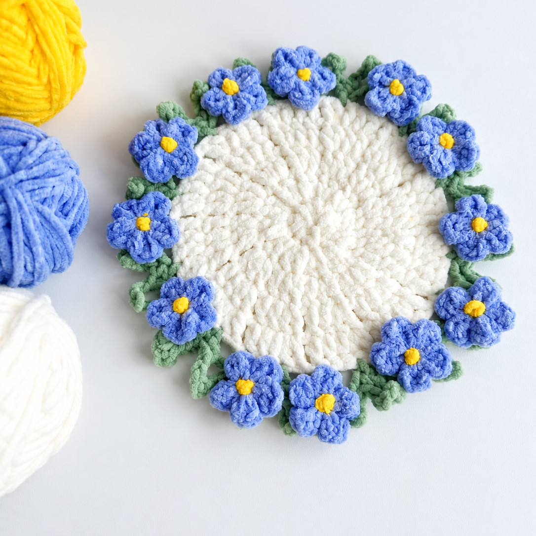 A white coaster in a soft plush yarn on a pale grey surface, ringed with green leaves and blue five-petal flowers with yellow centres, beside balls of yellow, blue and white yarn