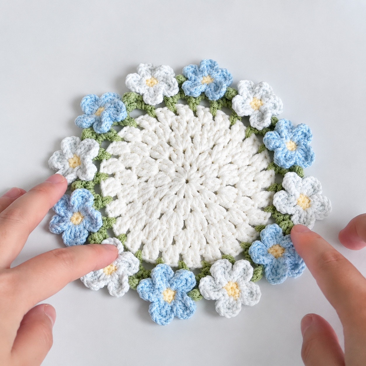 A forget-me-not coaster seen from above on a pale grey surface, held flat by two hands: a white openwork centre, a green leaf edging and five-petal flowers in blue and white with yellow centres