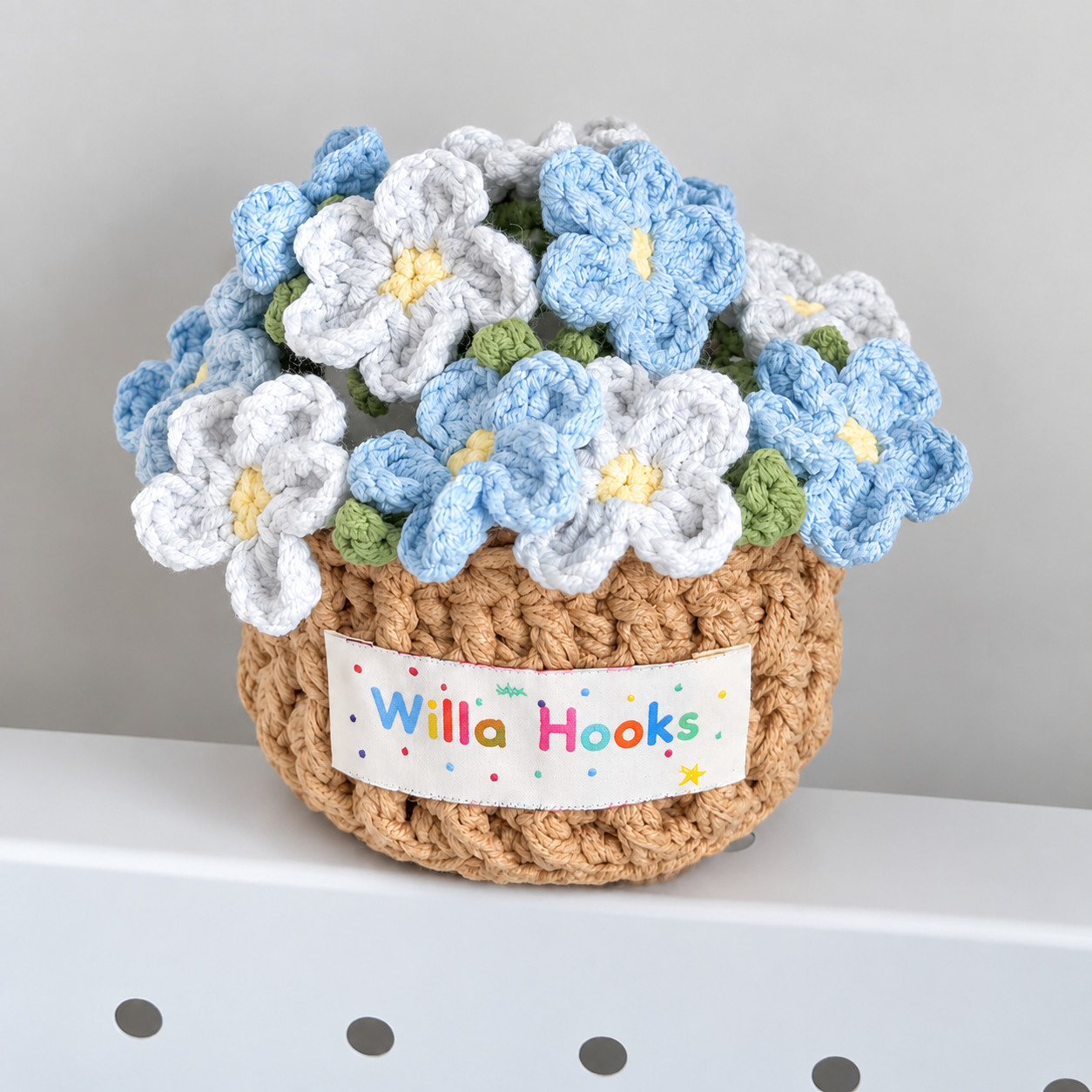 A tan ribbed crocheted flower pot with a Willa Hooks label, filled with blue and white crocheted forget-me-nots with yellow centres and small green leaves, on a white shelf