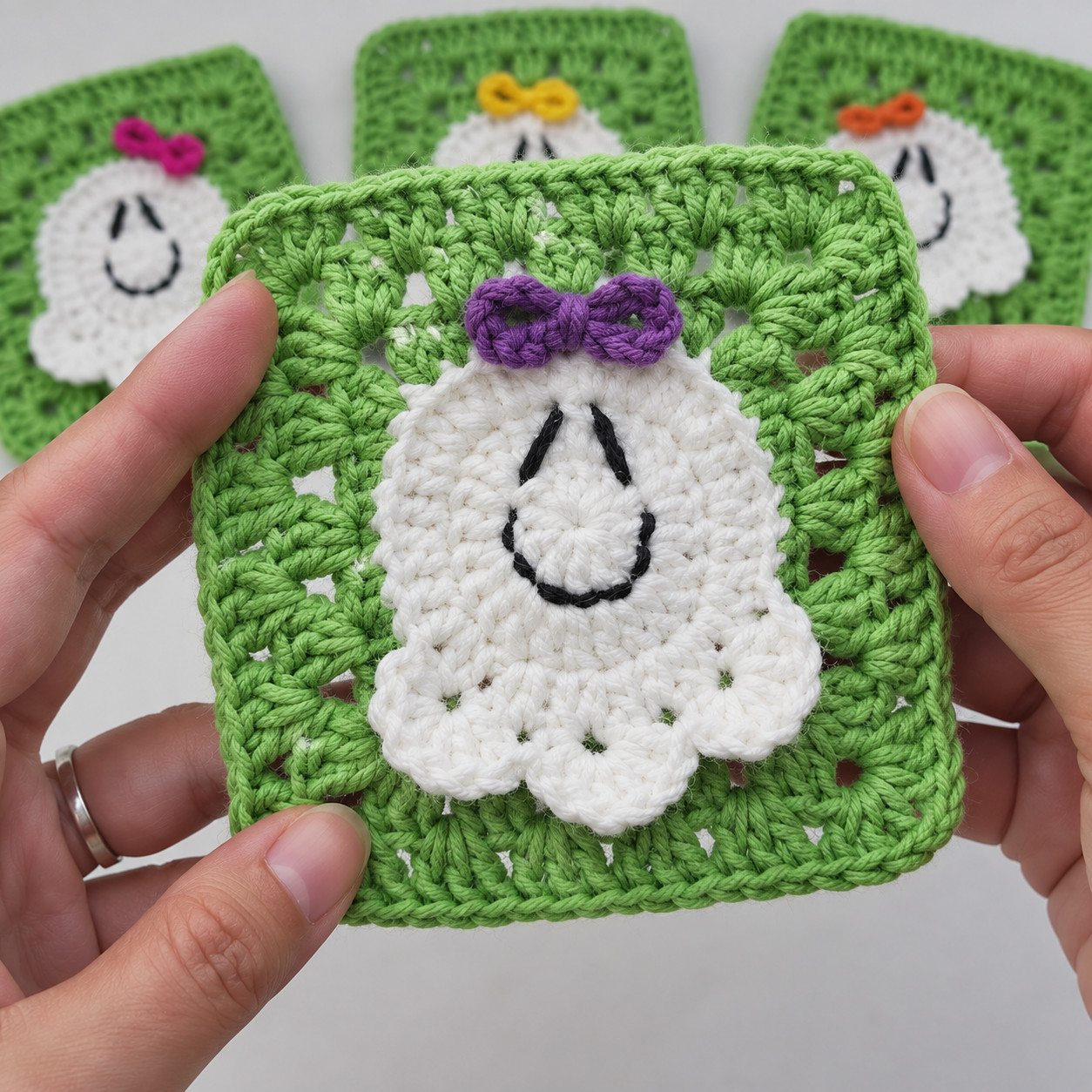 Two hands holding one green granny square with a white crocheted ghost and a purple bow, with three more ghost squares behind it
