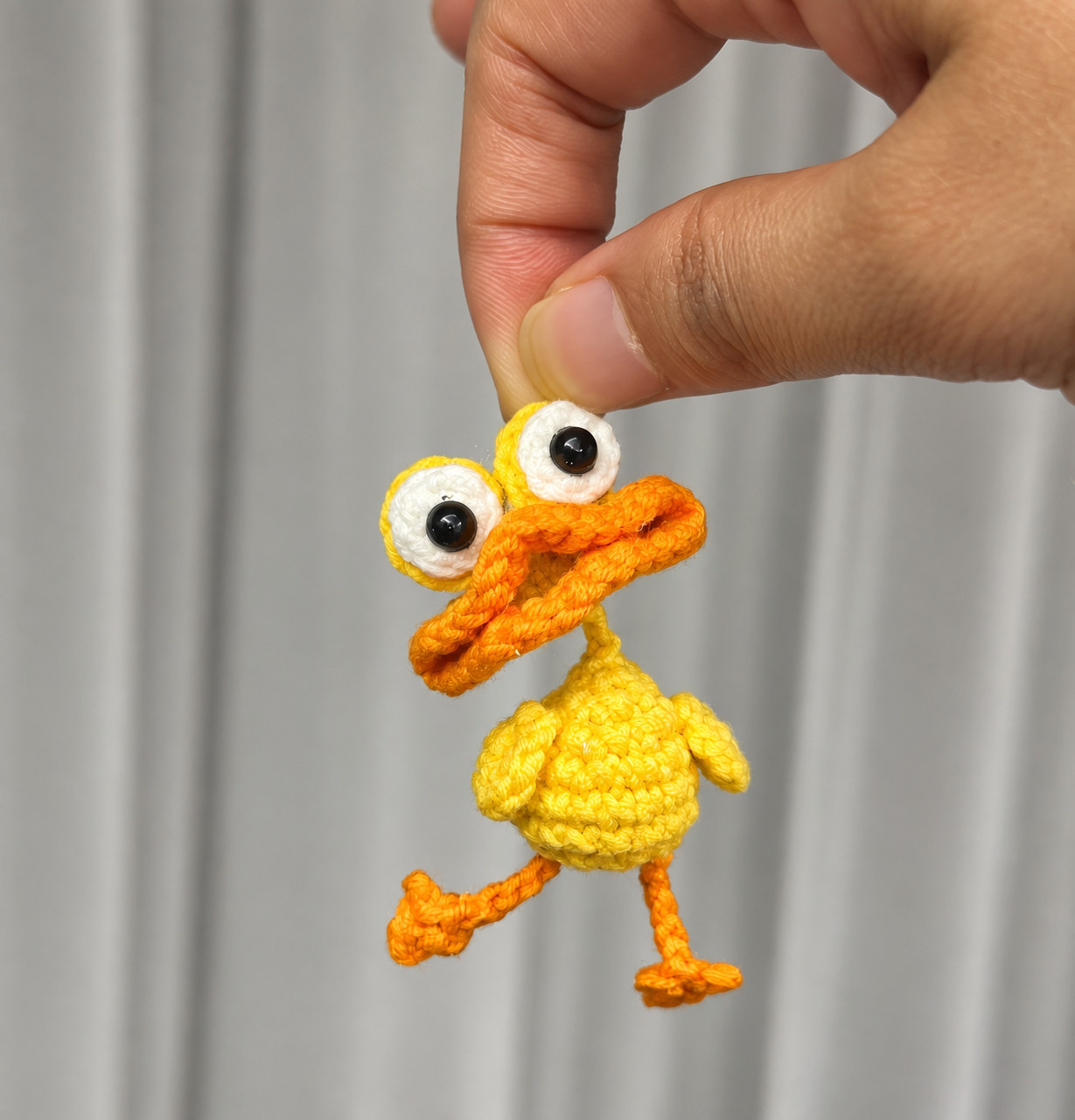 A crocheted yellow duck held up by the top of its head between finger and thumb in front of a grey curtain, with big white goggle eyes with black pupils, a wide folded orange bill, little yellow wings and long orange legs with fan-shaped feet, one kicked out to the side