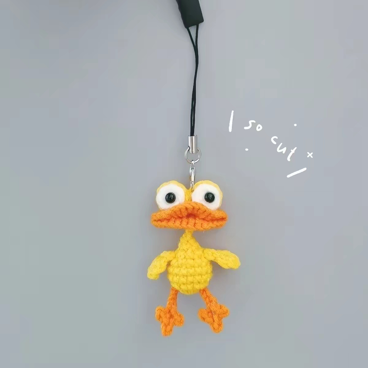 A crocheted yellow duck hanging from a black phone-strap cord on a small silver clasp against a grey background, with big white goggle eyes with black pupils, a wide orange bill, little yellow wings sticking out and two long orange legs with fan-shaped feet