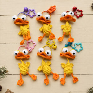 Six crocheted yellow ducks laid out in two rows on pale wooden boards, each with big white goggle eyes with black pupils, a wide folded orange bill, a round yellow body with little wings and long orange legs with fan-shaped feet; each hangs from a coloured ring with a star-shaped clasp and a bell, in purple, orange, red, pink, yellow and blue, with pine sprigs at the edges