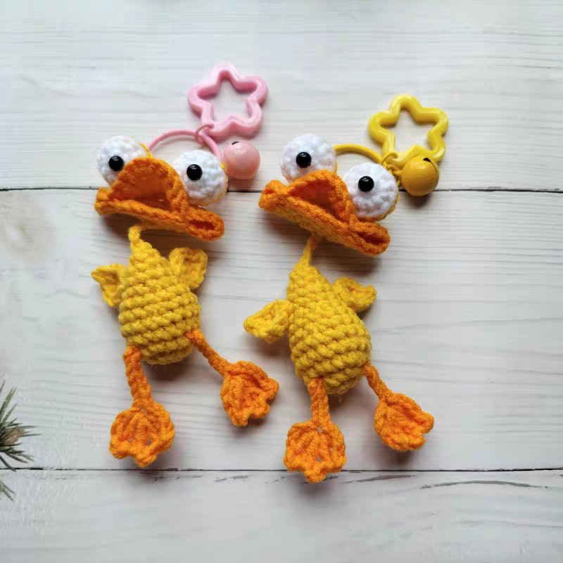 Two crocheted yellow ducks lying side by side on white wooden boards, each with big white goggle eyes with black pupils, a wide folded orange bill, a round yellow body with little wings and long orange legs with fan-shaped feet; one hangs from a pink ring with a pink star-shaped clasp and a bell, the other from a yellow ring with a yellow star-shaped clasp and a bell