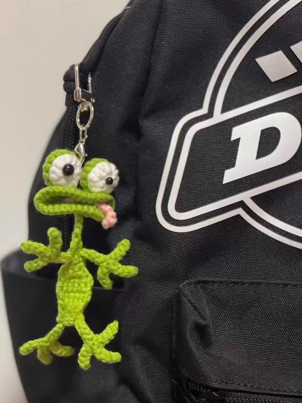 A crocheted green frog clipped by a silver clasp to a black backpack, with white goggle eyes, a pink tongue poking out of the corner of its wide mouth, and long skinny arms and legs with three toes each