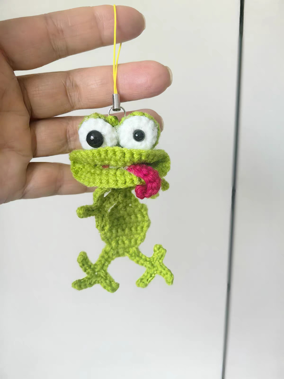 A crocheted green frog hanging from a yellow phone-strap cord held between two fingers, with white goggle eyes, one pupil bigger than the other, and a raspberry-red tongue curling out of its wide mouth