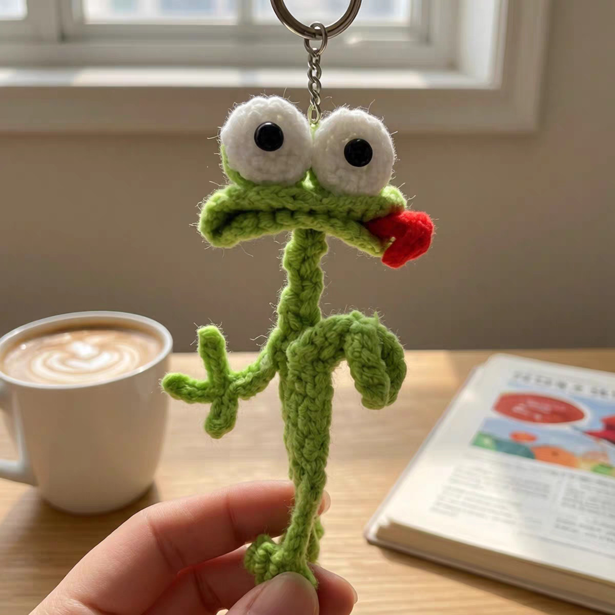 A crocheted green frog on a silver key ring and short chain, held up in the fingers in front of a bright window, with white goggle eyes, a red tongue poking out of its mouth and a long, skinny, twisted body; a cup of latte and an open book on the table behind