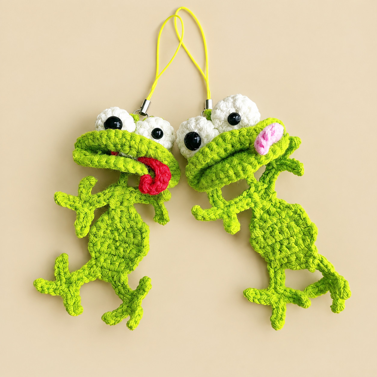 Two crocheted green frogs hanging side by side from yellow phone-strap cords on a beige background, each with big white goggle eyes, a wide folded mouth, a flat diamond body and long skinny arms and legs with three toes; one has a red tongue curling out of its mouth and the other a pink one