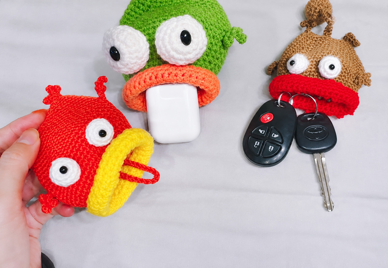 Three crocheted fish key pouches on a pale grey cloth: a red fish with a yellow lip held in a hand, a red cord looped out of its mouth; a green fish with an orange lip holding a white earbuds case; and a light brown fish with a red lip, two black car keys hanging from its mouth