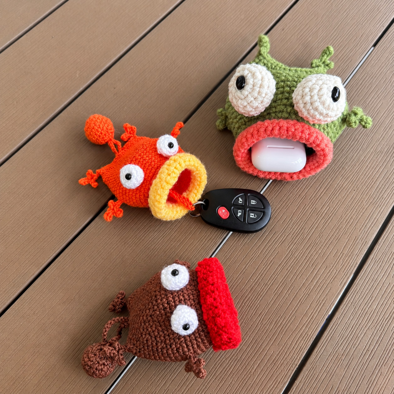 Three crocheted fish key pouches on wooden decking: an orange fish with a yellow lip and a bobble on its tail, a black car key fob hanging from its mouth; a green fish with a salmon-pink lip holding a white earbuds case; and a brown fish with a red lip lying on its side, its bobble on a cord behind it