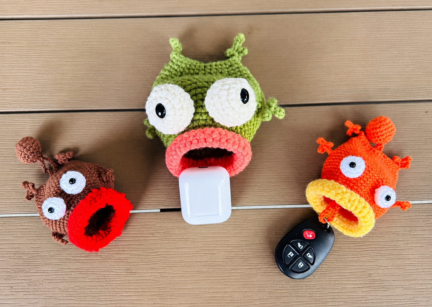 Three crocheted fish key pouches seen from above on wooden decking: a brown fish with a red lip and its mouth empty, a green fish with a salmon-pink lip with a white earbuds case in its mouth, and an orange fish with a yellow lip and a bobble on its tail, a black car key fob hanging below its mouth