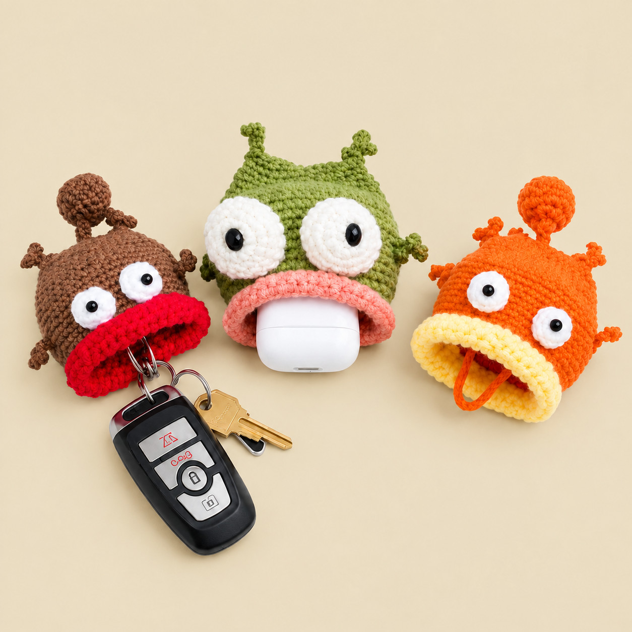 Three crocheted grumpy fish key pouches with white goggle eyes and thick rolled lips, on a cream background: a brown fish with a red lip, a car key fob and a house key hanging from its mouth; a green fish with a salmon-pink lip holding a white earbuds case; and an orange fish with a yellow lip, an orange cord hanging from its mouth and a bobble on its tail