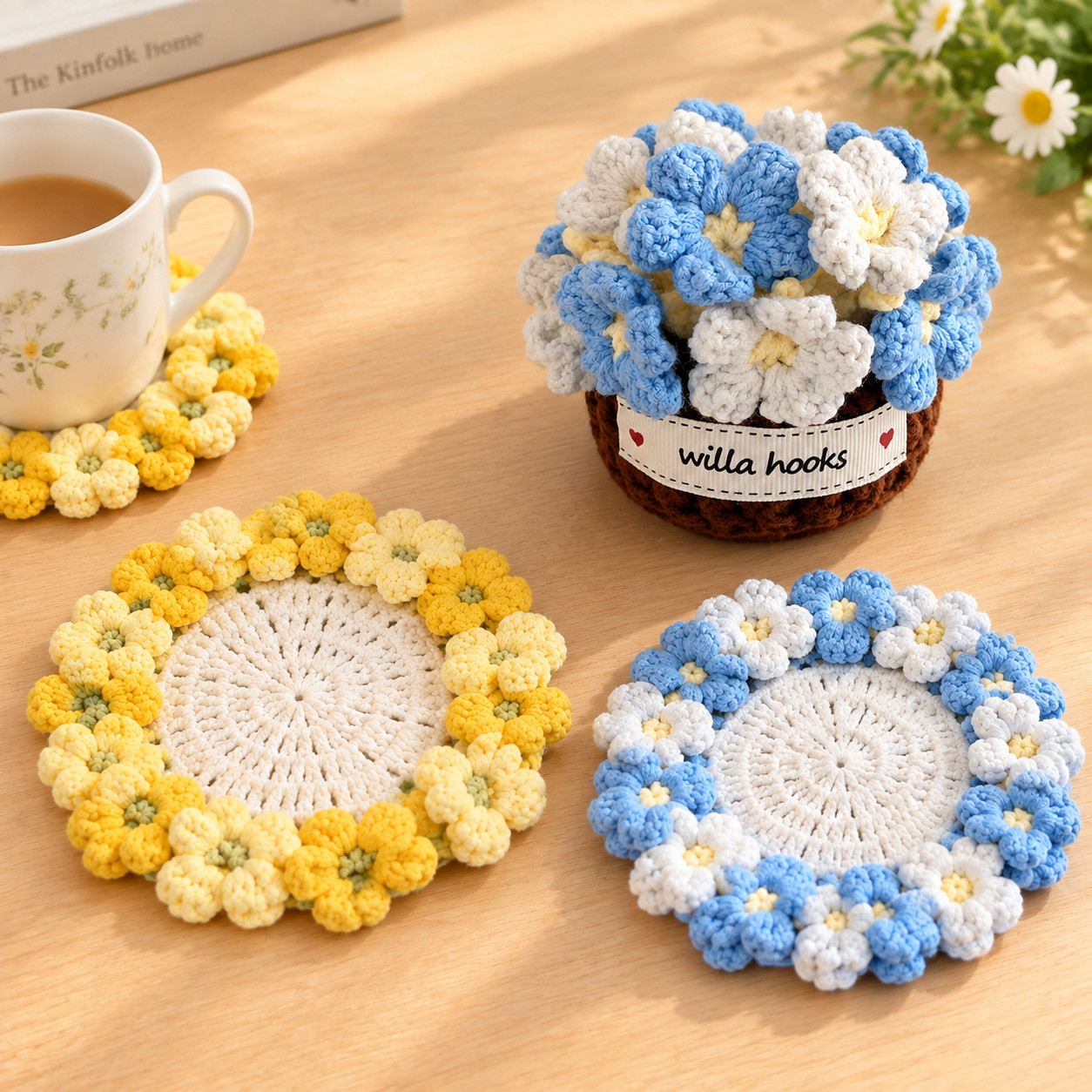 Two crocheted hydrangea coasters on a wooden table, one ringed with light and deep yellow flowers and one with blue and white flowers, beside a small dark brown crocheted pot with a Willa Hooks label holding a round head of blue and white flowers, and a third yellow coaster under a mug of tea