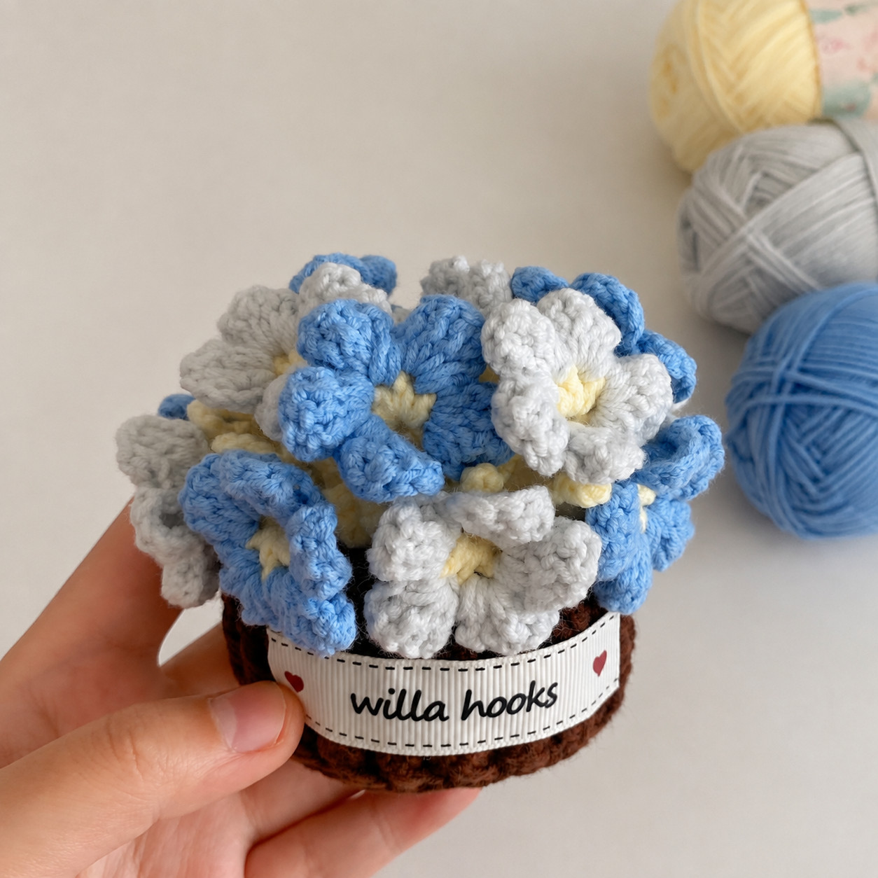 A hand holding a small dark brown crocheted pot with a Willa Hooks label, filled with a round head of blue and white crocheted flowers with pale yellow centres, beside balls of pale yellow, grey and blue yarn
