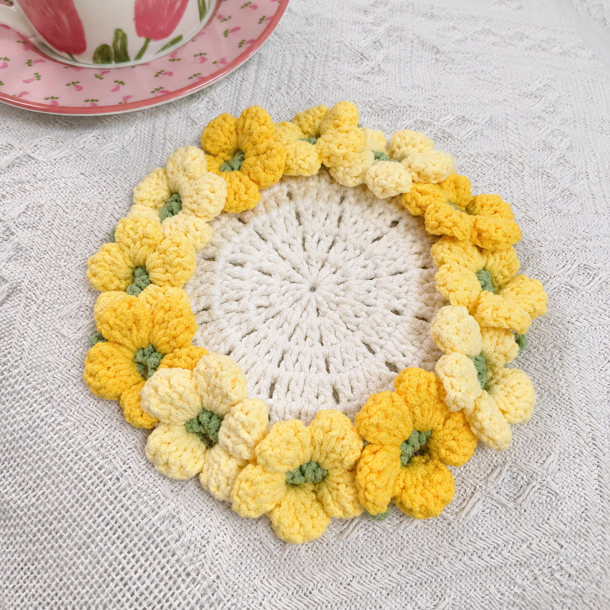 A crocheted hydrangea coaster on a white textured cloth: a cream openwork centre with lines of small gaps running out from the middle, ringed with light and deep yellow flowers with green centres, beside a teacup and saucer painted with pink tulips