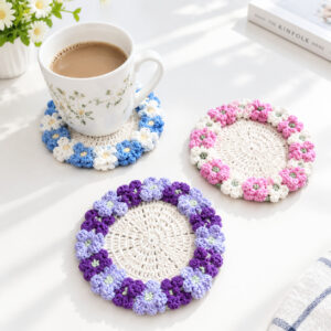 Three crocheted hydrangea coasters on a sunlit white table, each a cream openwork centre ringed with small flowers: blue and white under a mug of milky tea, pink and white, and purple and lilac