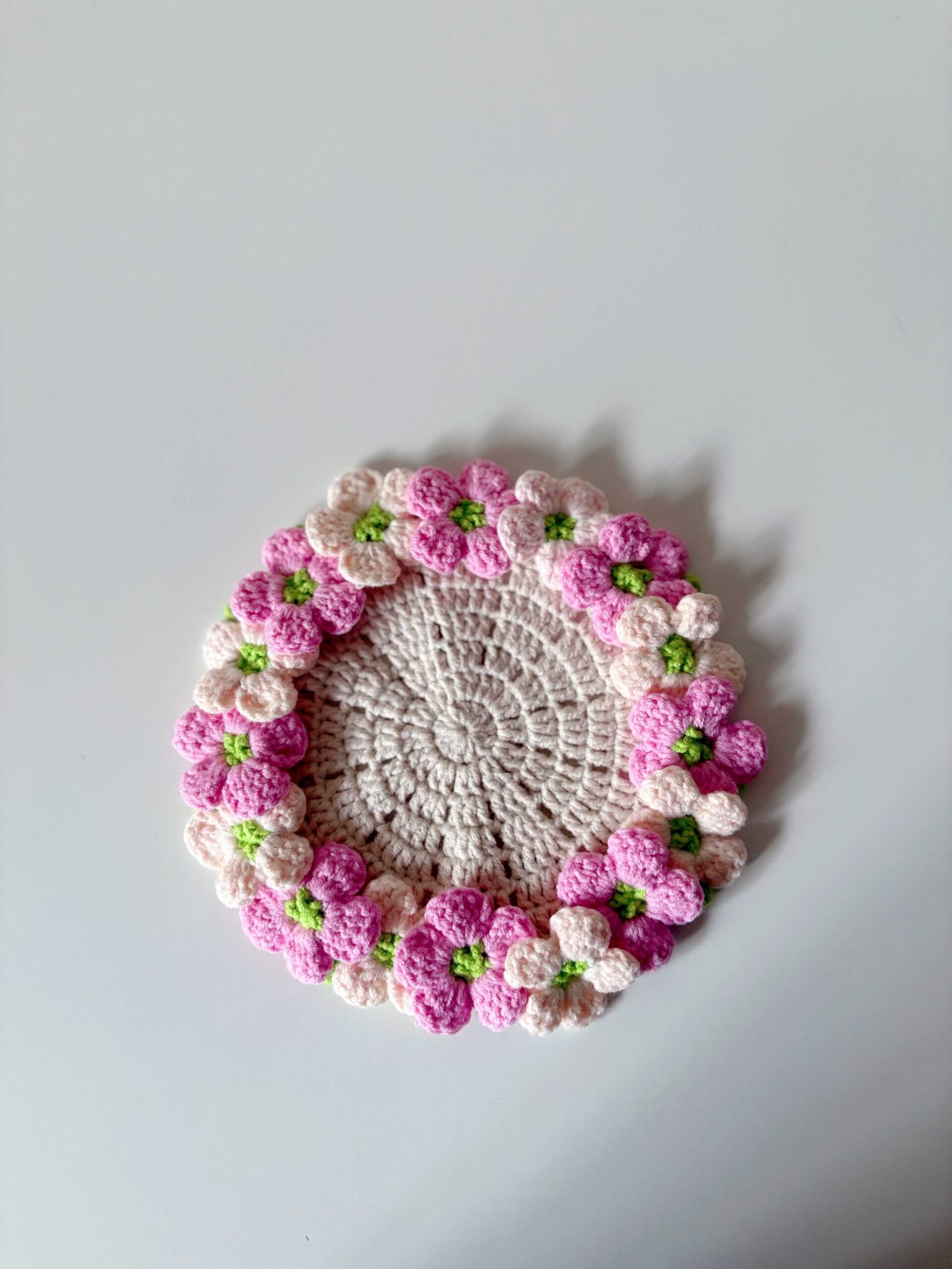 A crocheted hydrangea coaster seen from above on a white surface: a cream centre ringed with pink and pale pink flowers with bright green centres