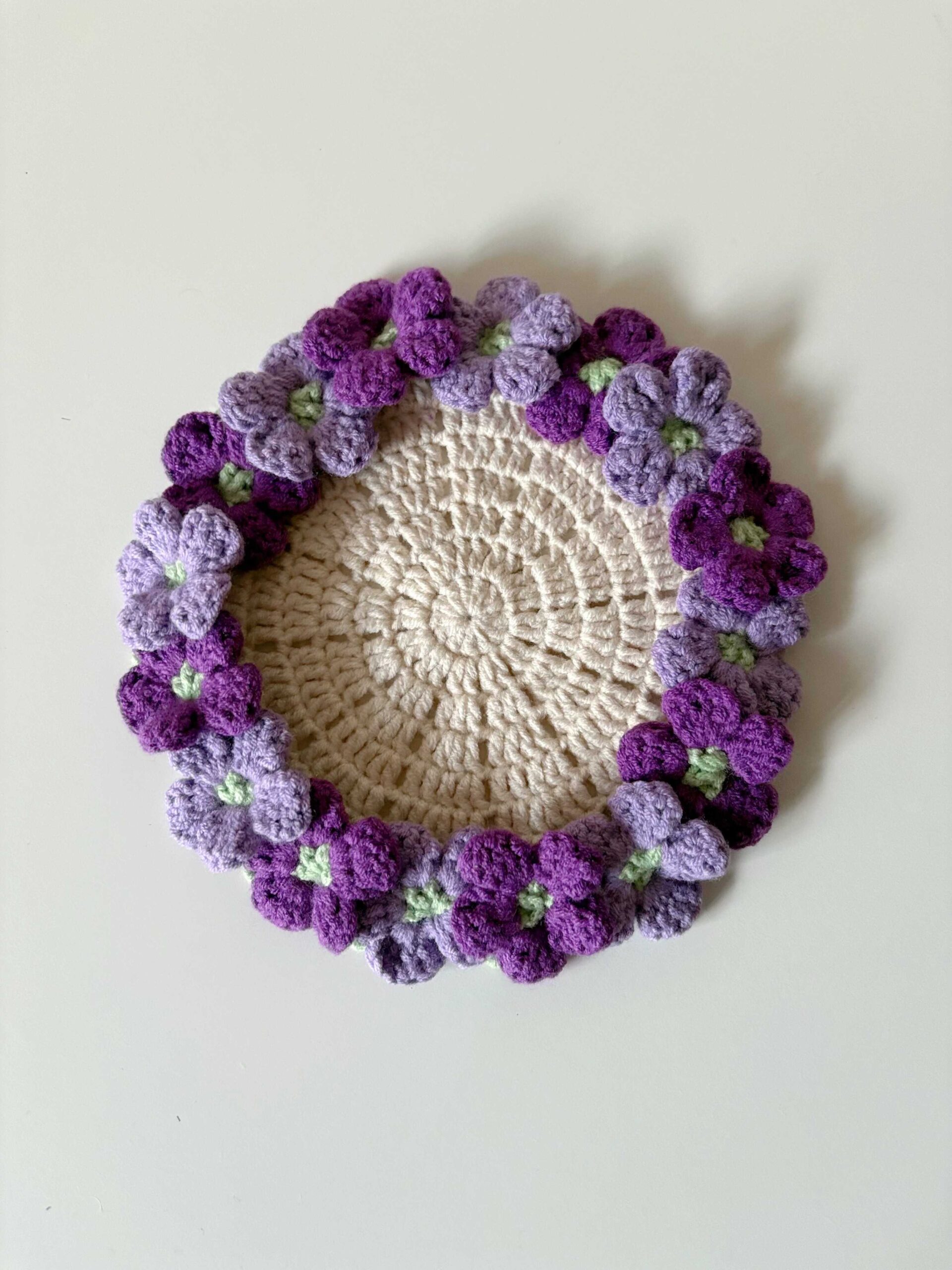 A crocheted hydrangea coaster seen from above on a white surface: a cream centre ringed with purple and lilac flowers with pale green centres