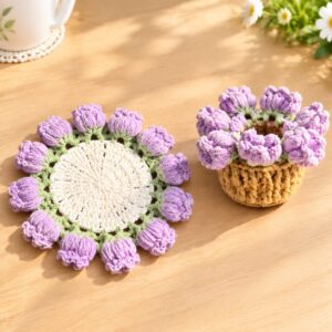 A crocheted lily of the valley coaster lying flat on a wooden table in sunlight: a cream centre, a pale green leaf edging and a ring of lilac bell-shaped lilies, beside a small mustard crocheted flower pot ringed with the same lilies