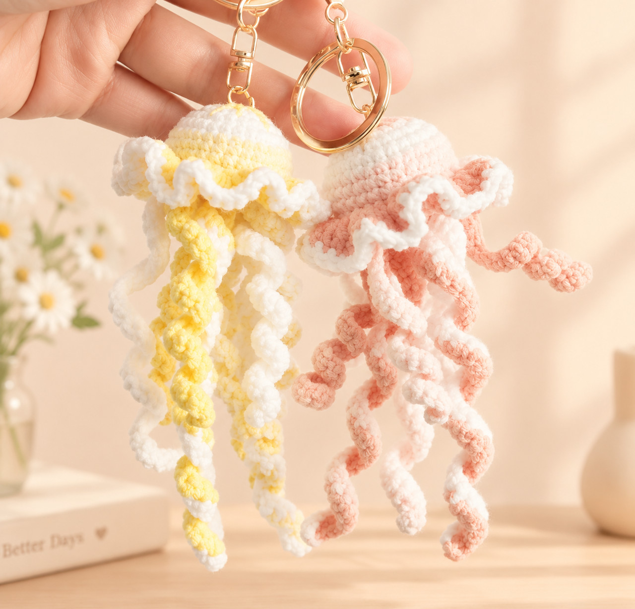 Two crocheted jellyfish keychains held up on gold key rings with swivel clasps, one yellow and white, one peach and white, their curly tentacles hanging down in front of a vase of daisies