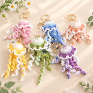 Six crocheted jellyfish keychains on gold key rings, laid on a cream surface among daisies: pink, blue, peach, yellow, green and purple, each with a white top, a ruffled skirt edged in white and curly tentacles