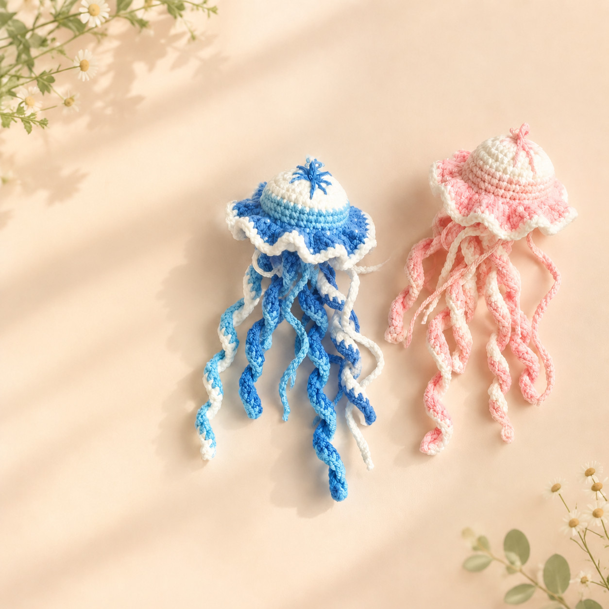 Two crocheted jellyfish lying on a peach surface in sunlight, one blue with lines stitched across its white top, one pink, each with a ruffled skirt and long curly tentacles