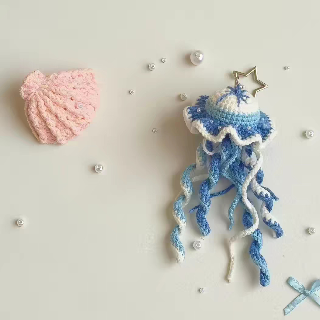 A blue and white crocheted jellyfish on a gold star-shaped key ring, with small pearls scattered around it and a pink crocheted shell beside it on a pale grey surface