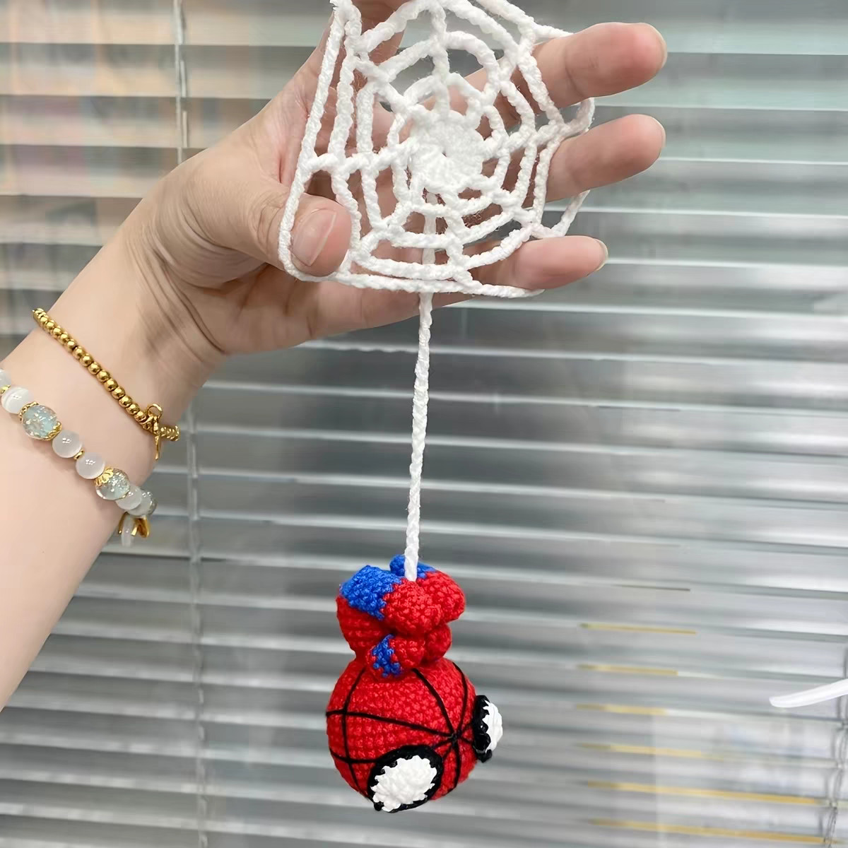 A round white crocheted web held flat in a hand in front of a window blind, a small red-and-blue crocheted figure hanging upside down beneath it on a white chain thread