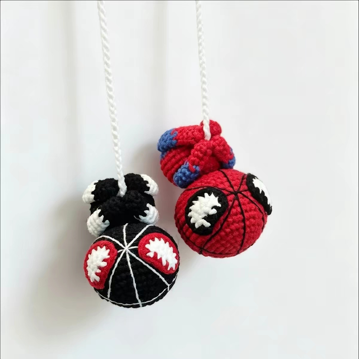Two small crocheted figures hanging upside down on white chain threads against a white wall: a black one with white lines across its head and white eyes edged in red, and a red-and-blue one with black lines across its head and white eyes edged in black