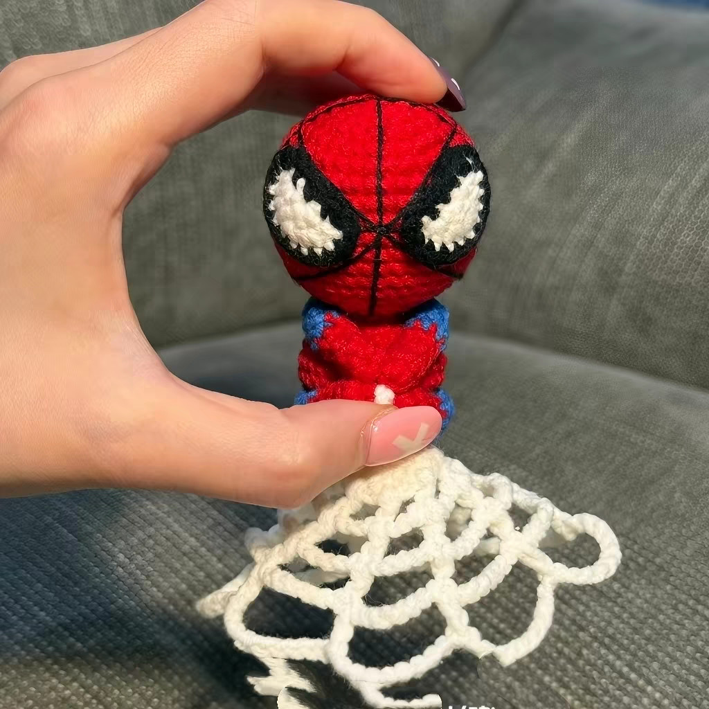 A small crocheted red-and-blue figure held between finger and thumb above a round white crocheted web on a grey sofa, its red head crossed with thin black lines and its big white eyes edged in black