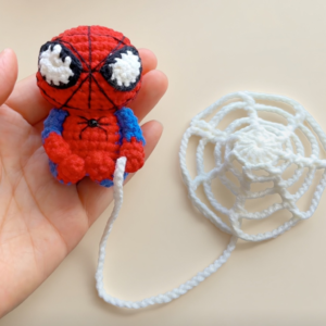 A small crocheted red-and-blue figure sitting in an open hand, with big white eyes edged in black, thin black lines across its red head and a small black spider on its chest, joined by a white chain thread to a round white crocheted web lying beside it